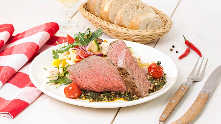 Ein Teller mit rosa gebratenem Fleisch, Gemüsebeilage, gebratenen Tomaten, Brot im Korb, Serviette und Besteck auf hellem Tisch.