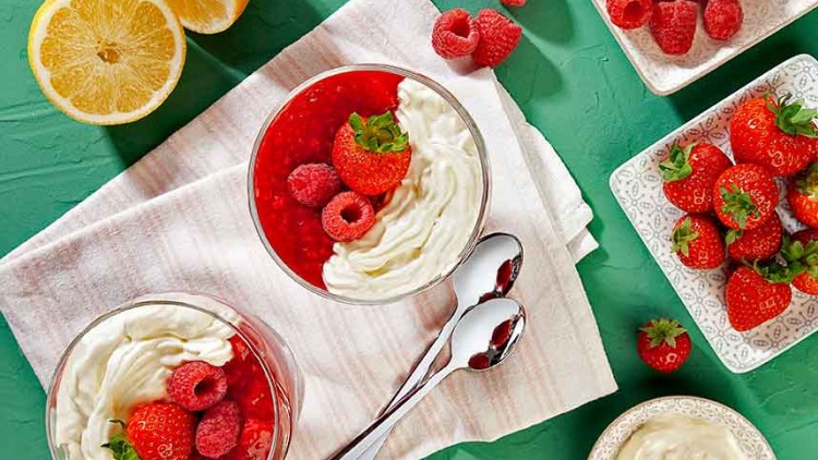 Zwei Dessertgläser mit roter Fruchtsoße, Sahne, Erdbeeren und Himbeeren auf Serviette, daneben Löffel und Schalen mit Früchten.