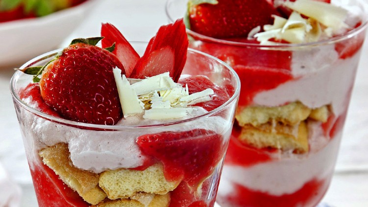 Zwei Gläser Schichtdessert mit Erdbeeren, weißer Schokolade, Sahnecreme und Biskuit auf hellem Untergrund.