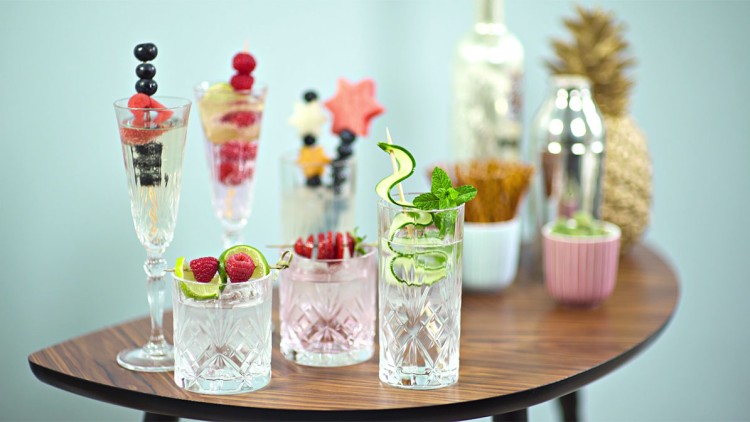 Fünf dekorierte Gläser mit Cocktails und Obst auf einem Holztisch, im Hintergrund Ananas, Flasche, Becher und Cocktailshaker.