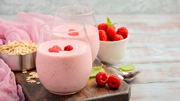 Zwei Gläser Himbeer-Smoothie mit frischen Himbeeren, Haferflocken in einer Schale und eine weiße Schale mit Himbeeren auf Holzbrett.