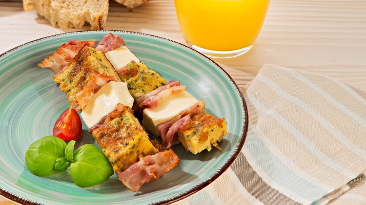 Grüner Teller mit Grillspießen aus Speck, Omelett und Käse, dekoriert mit Basilikum und Tomate, daneben Brot und Orangensaft.