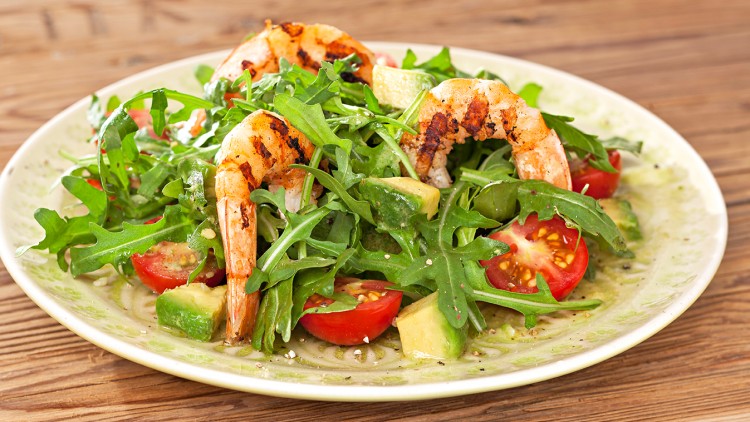 Ein Salatteller mit gegrillten Garnelen, Rucola, Avocado, Tomaten und Dressing auf einem Holztisch.