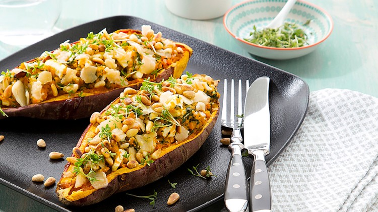 Zwei gefüllte Süßkartoffelhälften mit Käse, Kräutern und Nüssen auf schwarzem Teller, daneben Besteck und Stoffserviette.