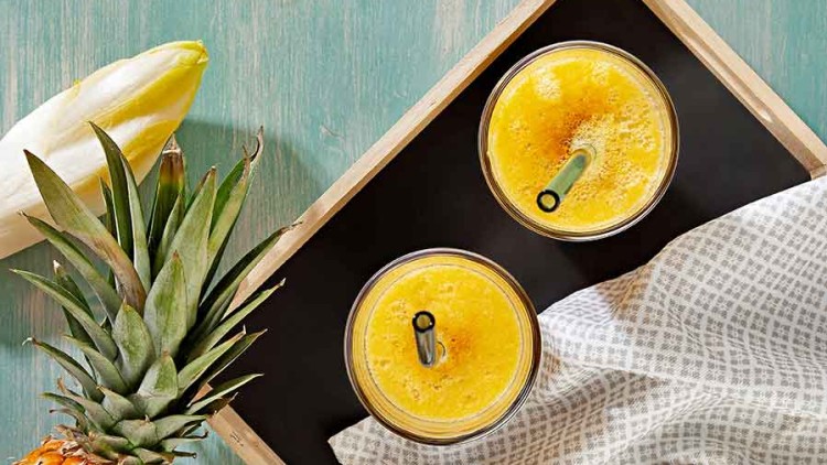Auf einem Tablett stehen zwei Gläser gelber Saft mit grünem Strohhalm, daneben Ananas, Chicorée, Ingwer und Kurkuma.