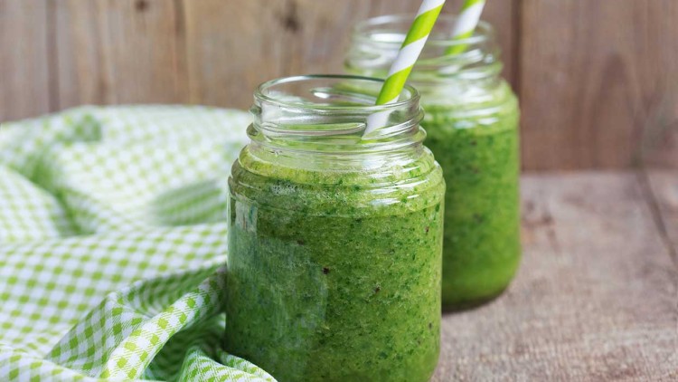 Zwei Gläser mit grünem Smoothie und gestreiften Trinkhalmen stehen vor einem Holz-Hintergrund auf einem Tisch mit karierter Serviette.