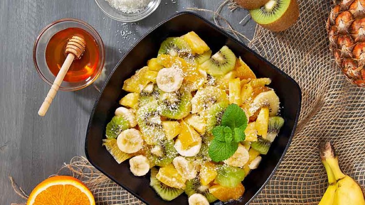 Schwarze Schale mit Obstsalat aus Banane, Kiwi, Orange und Ananas, dekoriert mit Minzblatt, daneben Honig und Kokosraspel.