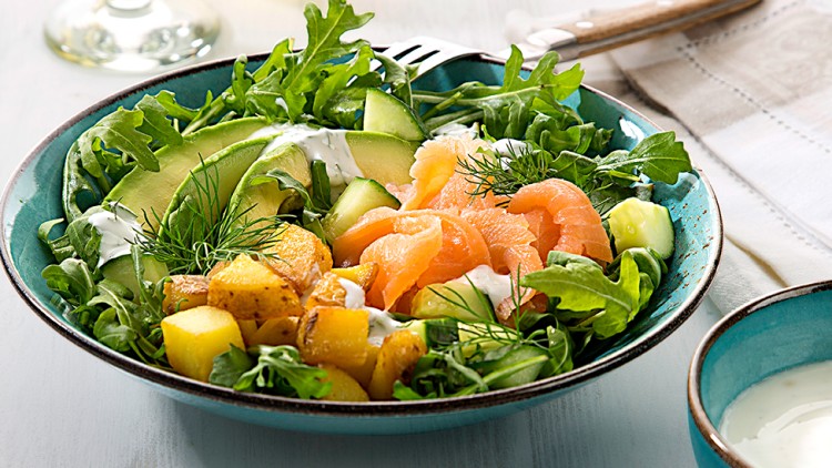 Schale mit grünem Salat, Gurke, Avocado, Lachs, Kartoffeln und Dill, daneben ein Glas Weißwein und ein Schälchen mit Sauce.