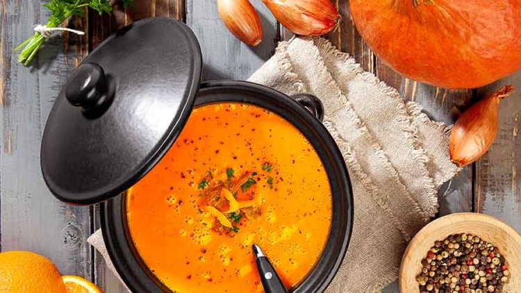 Eine Kürbissuppe in einem schwarzen Topf mit Löffel, daneben Kürbis, Schalotten, Pfeffer, Orange, Petersilie und ein Tuch auf Holz.