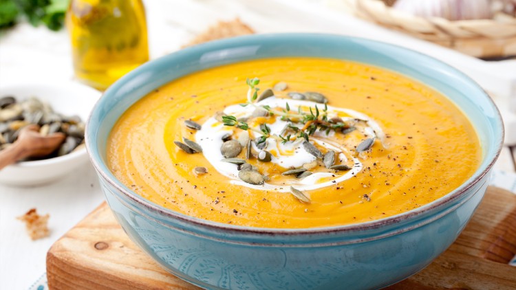 Orange Kürbissuppe in blauer Schüssel mit Sahne, Kürbiskernen, Thymian und Pfeffer garniert auf Holzbrett serviert.