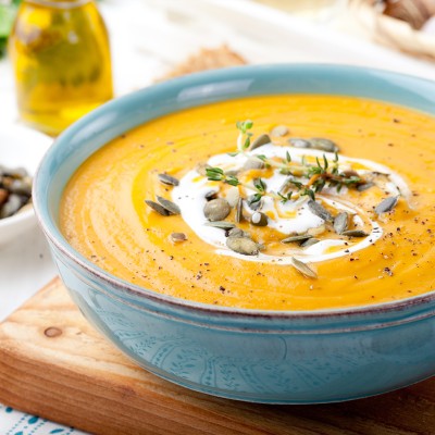 Orange Kürbissuppe in blauer Schüssel mit Sahne, Kürbiskernen, Thymian und Pfeffer garniert auf Holzbrett serviert.