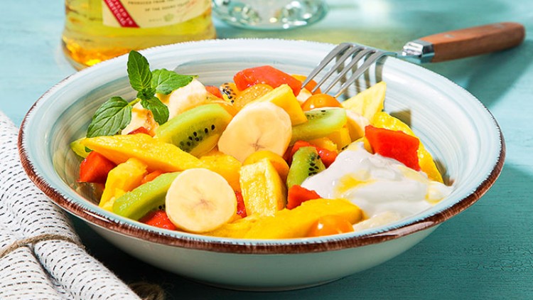 Schale mit Obstsalat aus Bananen, Kiwi, Papaya und Mango sowie Joghurt, garniert mit Minzblatt und Besteck auf Holzuntergrund.