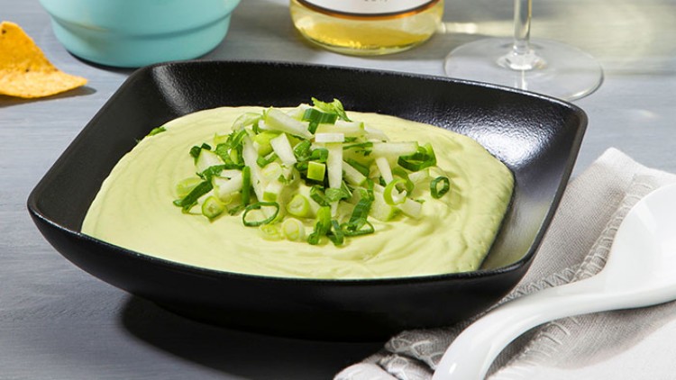Eine schwarze Schale mit grüner Creme, garniert mit Frühlingszwiebeln, steht neben Löffel, Serviette, Chips und einer Weinflasche.