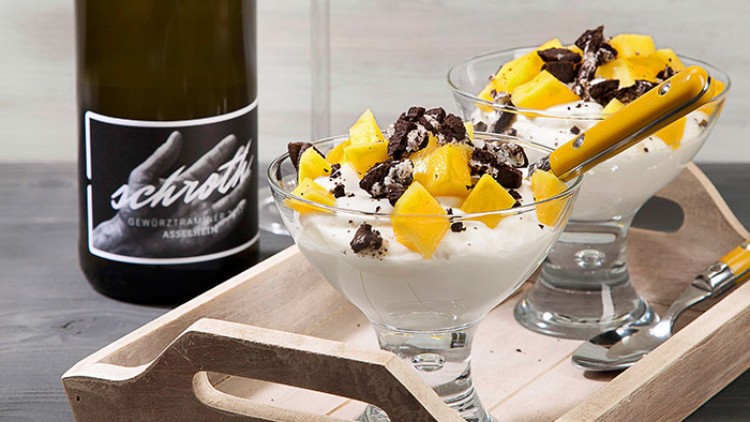 Zwei Dessertgläser mit Joghurt, Mango und Schokostücken auf einem Holztablett, daneben eine Weinflasche mit dem Schriftzug „schrotty“.