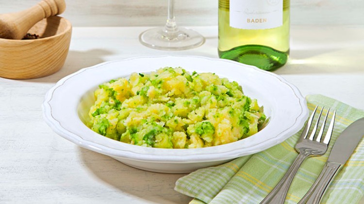 Weiße Schüssel mit Kartoffelsalat, daneben ein Glas Weißwein und eine Flasche Gutedel, Messer und Gabel liegen auf grünem Tuch.