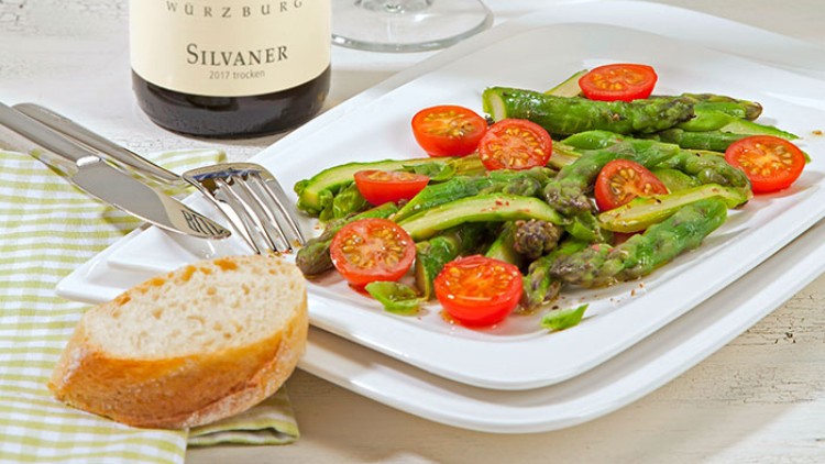 Grüner Spargel mit halbierten Kirschtomaten auf einem weißen Teller, daneben Weißbrot, Besteck und eine Flasche Silvaner.