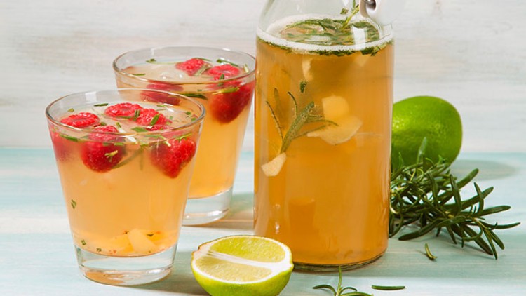 Eine Glasflasche und zwei Gläser mit gelbem Getränk, Himbeeren, Limette, Rosmarin und Eiswürfeln auf hellem Untergrund.