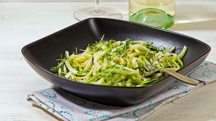 Ein Teller Zucchini-Spaghetti mit frischen Kräutern, serviert in einer schwarzen Schale, daneben ein Glas und eine Flasche Weißwein.