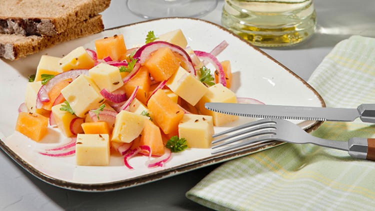 Würfel aus Käse und Melone, rote Zwiebeln und Petersilie auf einem Teller, daneben Brotscheiben, Besteck, ein Weinglas und eine Flasche.