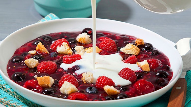 Rote Beerengrütze mit Himbeeren und Heidelbeeren in einem weißen Teller, übergossen mit Sahne aus einem Glaskrug.