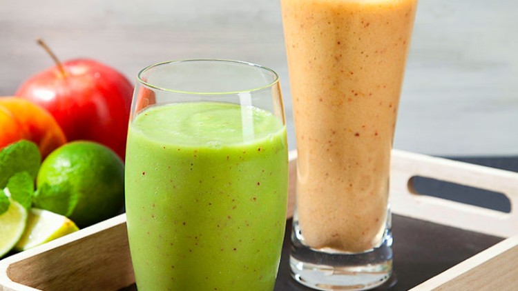 Ein grüner und ein orangefarbener Smoothie in Gläsern auf einem Holztablett, daneben Äpfel, Limetten und Minzblätter.