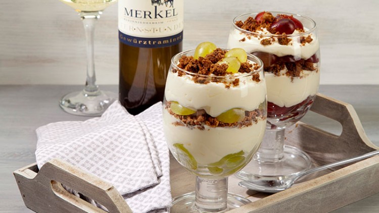 Zwei Dessertgläser mit Cremeschichten, Trauben und Bröseln auf Holztablett, daneben eine Flasche Weißwein und ein Glas.