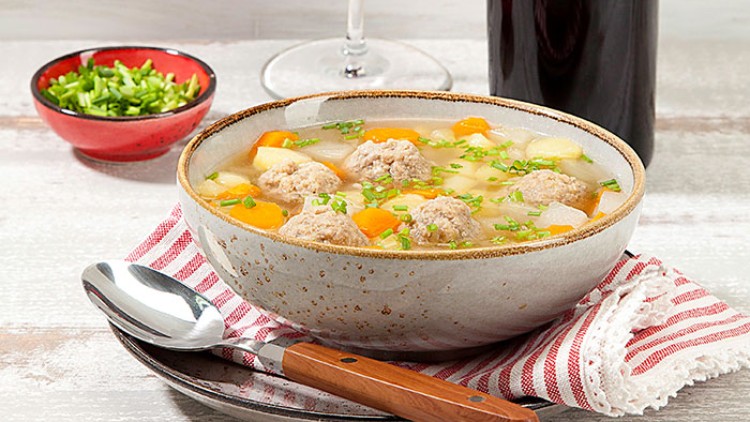 Keramische Schale mit Gemüsesuppe und Fleischklößchen auf gestreiftem Stofftuch, daneben ein Löffel und gehackte Kräuter.