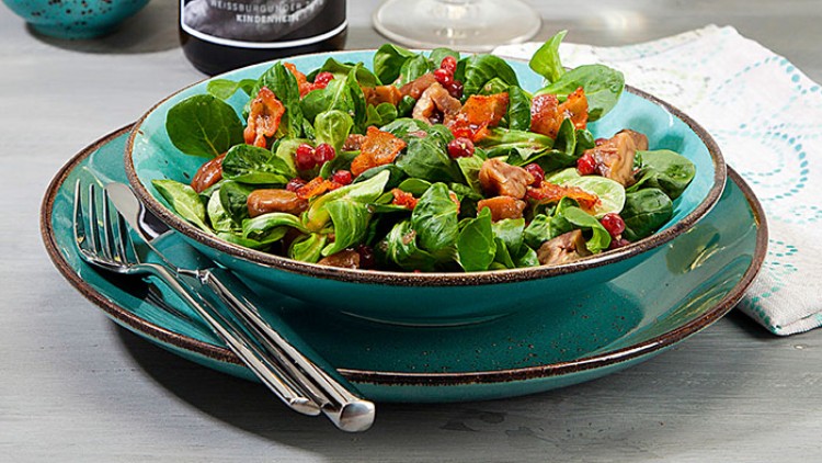 Ein Teller mit grünem Feldsalat, Pilzen, roten Beeren und Speck auf einem Stapel türkiser Keramik, daneben Besteck.
