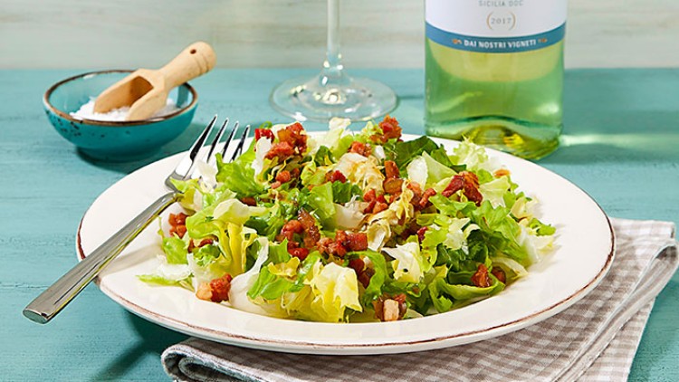 Ein Teller mit bunter Blattsalatmischung und Speckwürfeln, daneben ein Glas Weißwein und eine Flasche mit der Aufschrift „Grillo“.