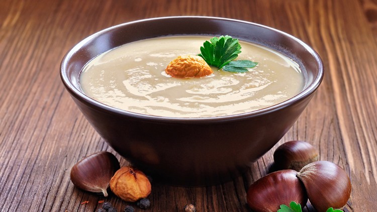 Braune Schale mit Kastaniensuppe, garniert mit einer ganzen Kastanie und Petersilie, umgeben von Kastanien und Gewürzen auf Holz.