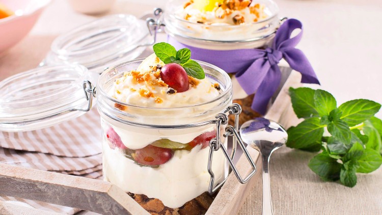 Zwei gefüllte Einmachgläser mit Schichtdessert aus Creme, Obst und Streuseln, eines dekoriert mit Minze und Kirsche.