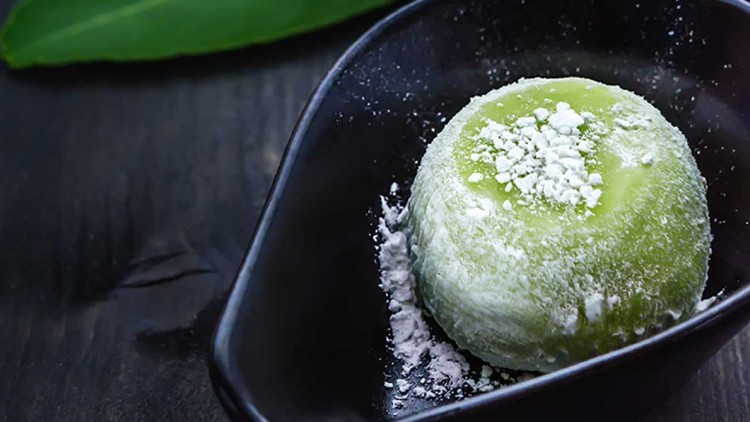 Ein grünes Mochi mit Puderzucker liegt in einer schwarzen Schale, daneben dampft eine schwarze Tasse und grüne Blätter sind sichtbar.