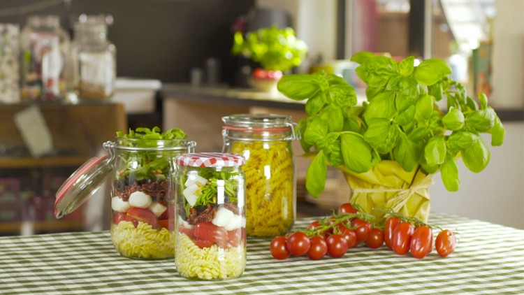Drei Gläser mit Nudelsalat und Kräutern, frische Basilikumpflanze und Strauchtomaten auf einem grün-weiß karierten Tischtuch.