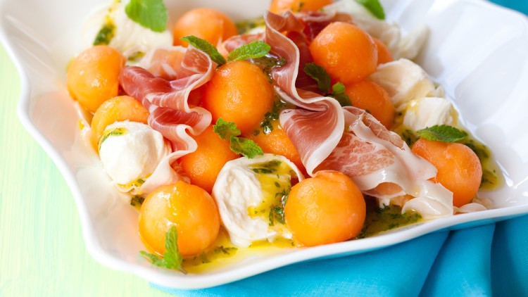 Kugeln aus orangefarbener Melone, Mozzarella, Schinkenstreifen und Minzblätter auf weißem Teller mit Dressing.