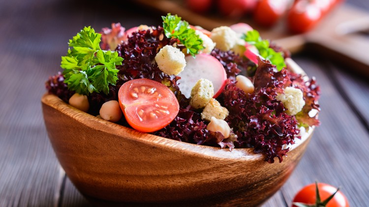 Roter Blattsalat mit Petersilie, Kichererbsen, Tomaten, Radieschen und Croutons in einer Holzschale auf dunkler Holzoberfläche.