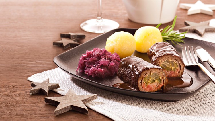 Schwarzer Teller mit Rotkohl, zwei Rouladen und zwei Kartoffelklößen, daneben Besteck und Holzsterne auf Holzuntergrund.