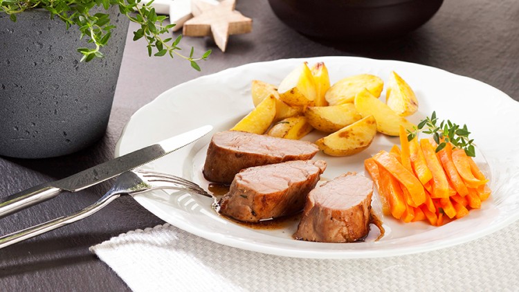 Weißer Teller mit Schweinefilet, Kartoffelspalten und Möhren, daneben Kräutertöpfe und Holzdekoration in Stern- und Baumform.