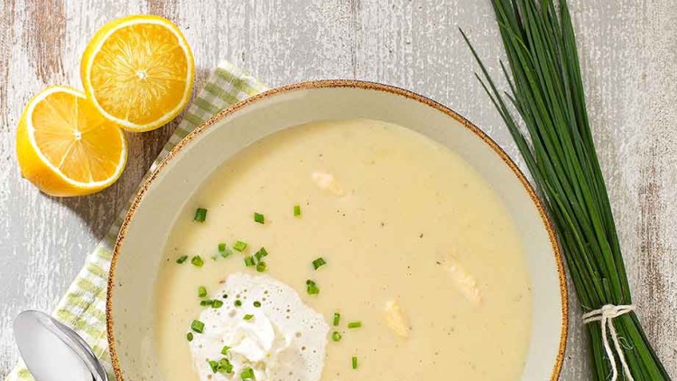 Ein Teller mit heller Suppe, Sahnehäubchen und Schnittlauch, daneben Schnittlauchbündel, Schalotten, Zitrone und Löffel auf kariertem Tuch.