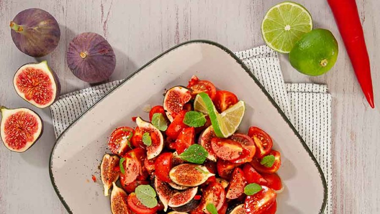 Quadratischer Teller mit Salat aus geschnittenen Feigen, Tomaten, Pfeffer und Minze, dekoriert mit Limettenvierteln, auf Tisch.