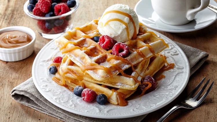 Belgische Waffeln mit Karamellsoße, Eis, Himbeeren und Blaubeeren auf einem weißen Teller, daneben Kaffee und Schale mit Beeren.