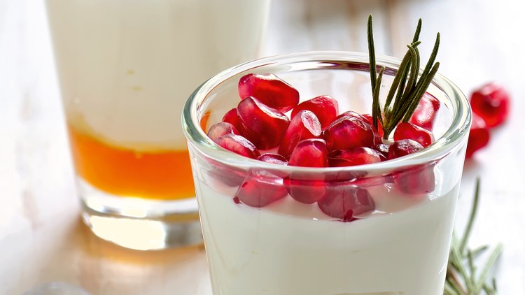 Ein Glas mit weißem Joghurt, Granatapfelkernen und einem Zweig Rosmarin, im Hintergrund ein weiteres Schichtdessert.