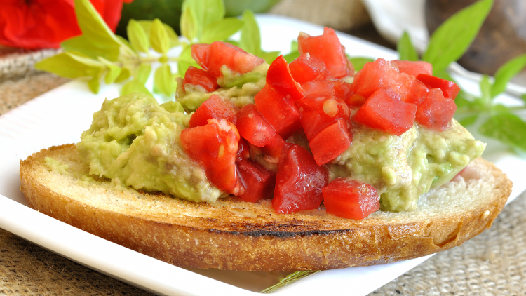 Getoastete Brotscheibe mit Guacamole und frischen Tomatenwürfeln, dekoriert mit grünen Kräutern auf weißem Teller.