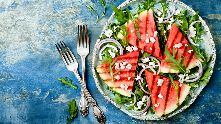 Ein Teller mit Rucola, roten Zwiebelringen, gegrillten Wassermelonen-Stücken und zerbröseltem weißen Käse, daneben zwei Gabeln.
