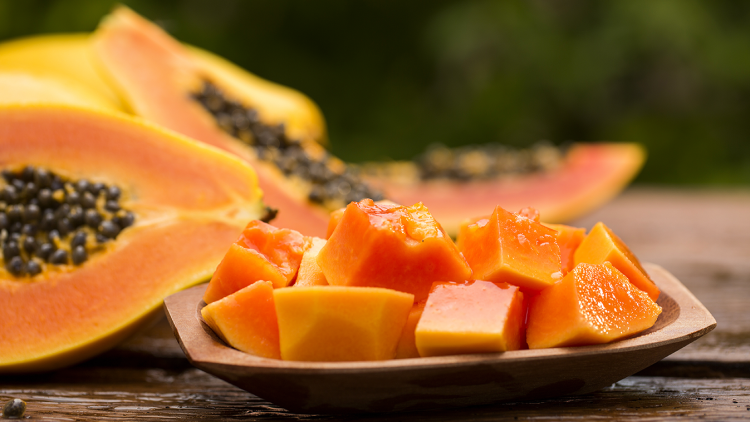 Holzschale mit orangefarbenen Papayastücken, im Hintergrund halbierte Papayas mit schwarzen Kernen auf Holztisch.