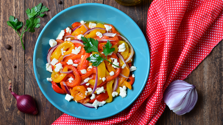 Bunter Salat aus Paprikastreifen, roten Zwiebeln, Fetastücken und Petersilie in einer blauen Schale, daneben Öl und rote Zwiebel.