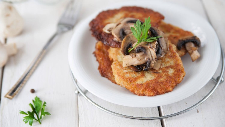 Zwei goldbraune Kartoffelpuffer mit gebratenen Champignons und frischer Petersilie auf einem weißen Teller.