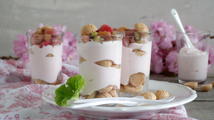 Drei Gläser mit Schichtdessert aus rosa Creme, Keksen und Obststücken auf einem weißen Teller mit frischer Minze.