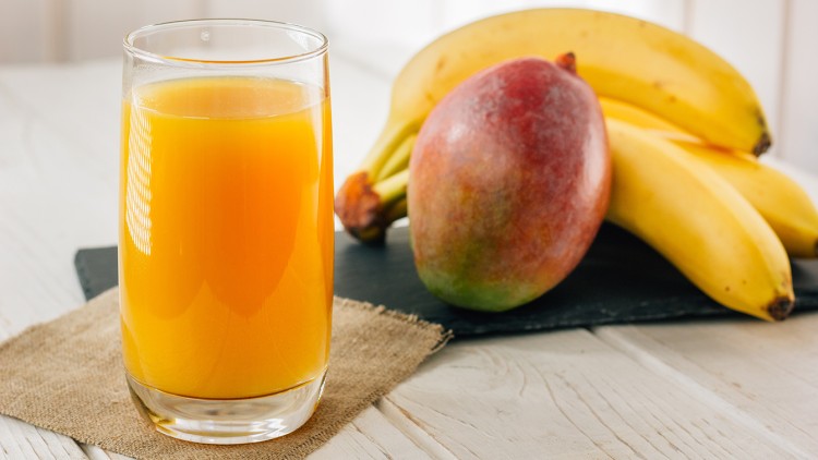 Ein Glas mit orangefarbenem Saft steht auf einem hellen Holztisch vor einer Mango und mehreren Bananen auf einer Schieferplatte.