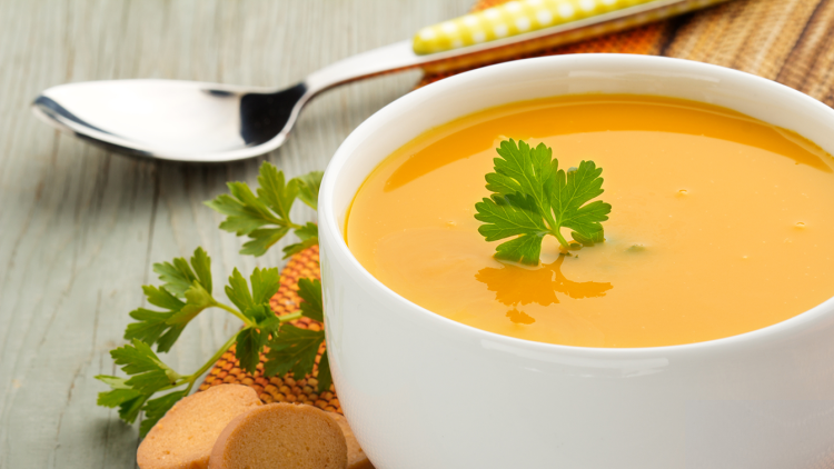 Weiße Schale mit orangefarbener Suppe, dekoriert mit Petersilie, daneben Brotchips, Petersilie und ein Löffel mit gelbem Griff.