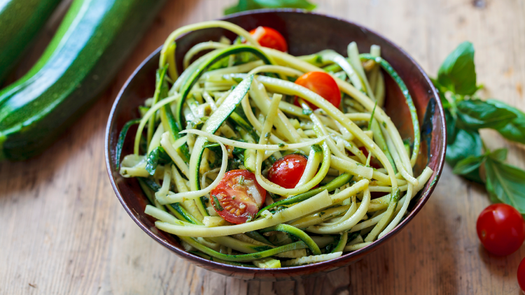 Schüssel mit Zucchininudeln, kleinen Tomaten und Kräutern, daneben frische Zucchini, Basilikum und Kirschtomaten auf Holz.